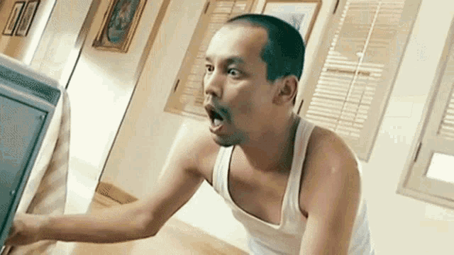 a man in a white tank top is sitting in front of a television with his mouth open