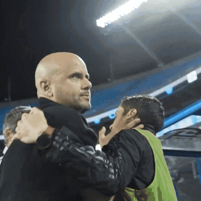 a bald man is hugging another man in a green vest