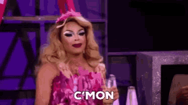 a drag queen in a pink dress is holding a glass of champagne and saying c'mon .