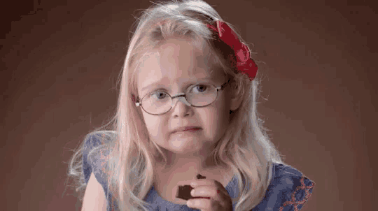 a little girl wearing glasses is eating a piece of chocolate and making a sad face .
