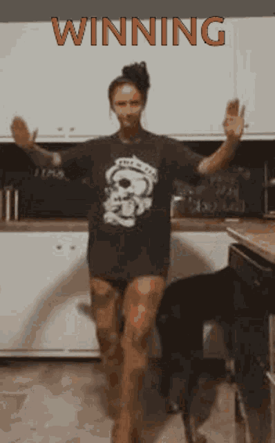 a woman in a skull shirt is dancing in a kitchen with the word winning above her