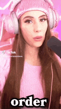 a woman wearing headphones and a pink sweater says order in front of a microphone