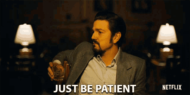 a man in a suit is holding a glass of whiskey and says " just be patient "