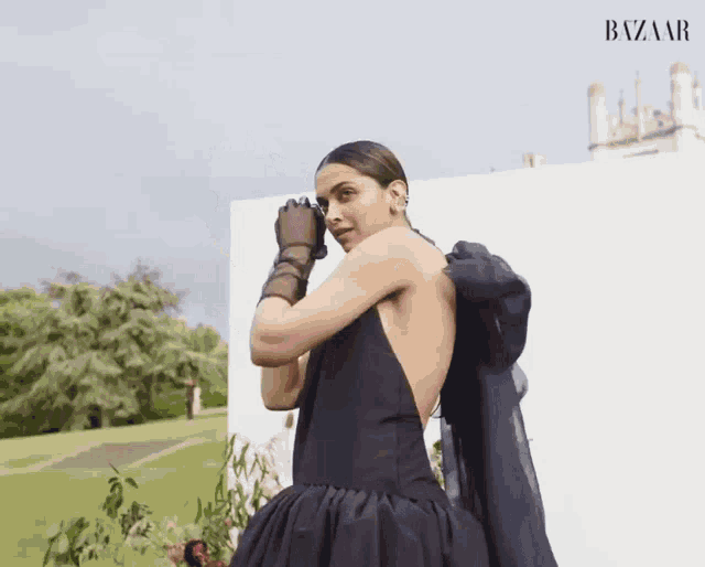 a woman in a black dress and black gloves is standing in front of a white wall that says bazaar on it