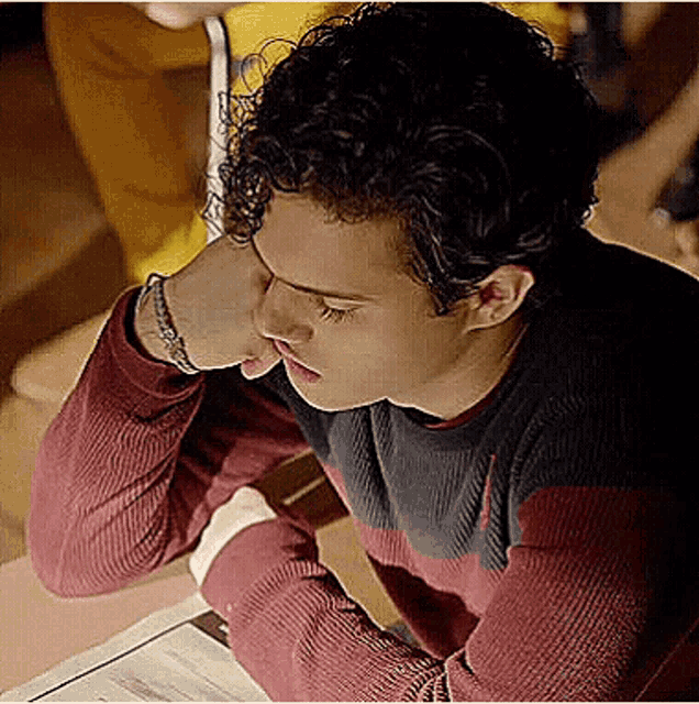 a young man with curly hair is sitting at a table with his hand on his face .