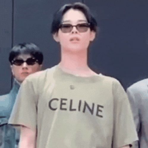 a man wearing a green celine t-shirt and sunglasses is standing in front of a group of people .