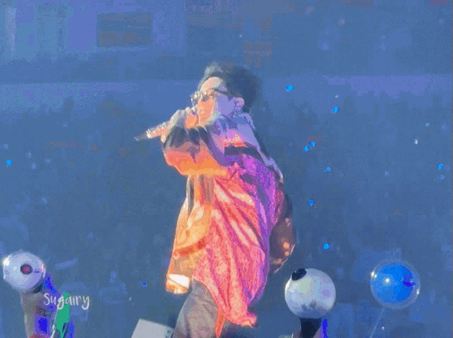 a man in an orange shirt is holding a microphone in front of a crowd with sugary written on the bottom