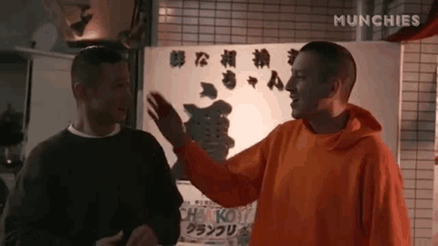 two men in orange hoodies are standing next to each other in front of a sign .