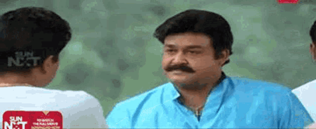 a man with a mustache wearing a blue shirt is talking to another man .