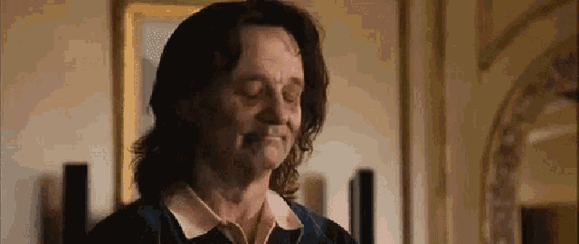 a man with long hair is standing in a room with his eyes closed and smiling .