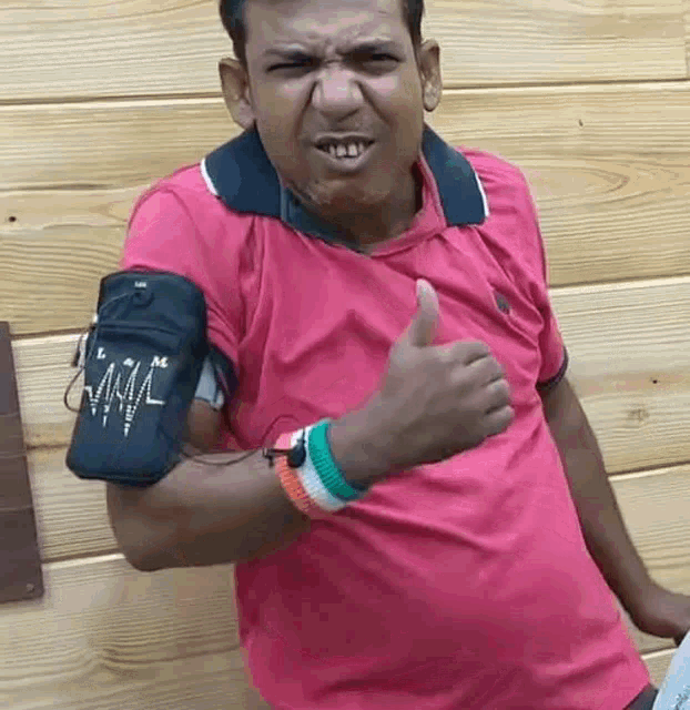 a man in a pink shirt is giving a thumbs up sign while standing in front of a wooden wall .