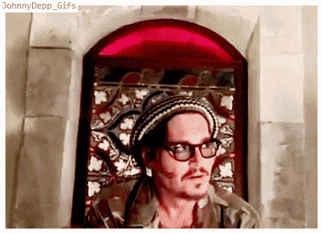 a man wearing glasses and a knitted hat stands in front of a window with the caption johnny depp gifs