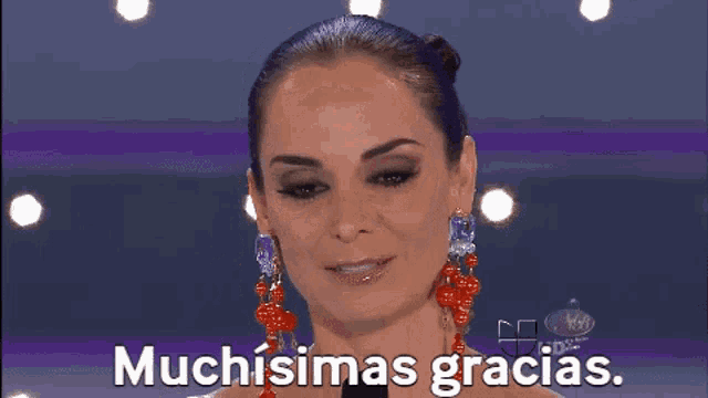 a woman wearing earrings and a bun is smiling and says muchisimas gracias