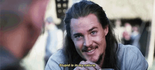 a man with long hair and a beard is talking to another man while holding a glass .