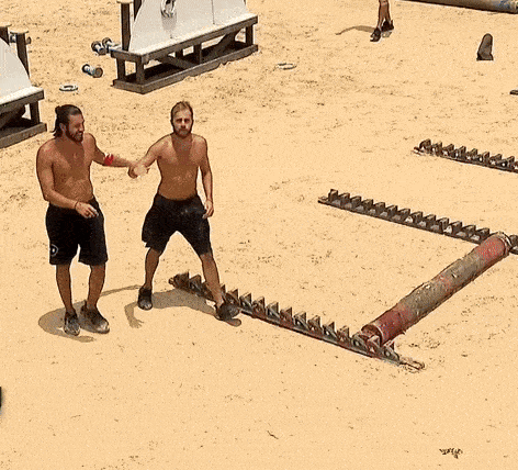 two shirtless men are standing on a sandy beach and one of them is pointing