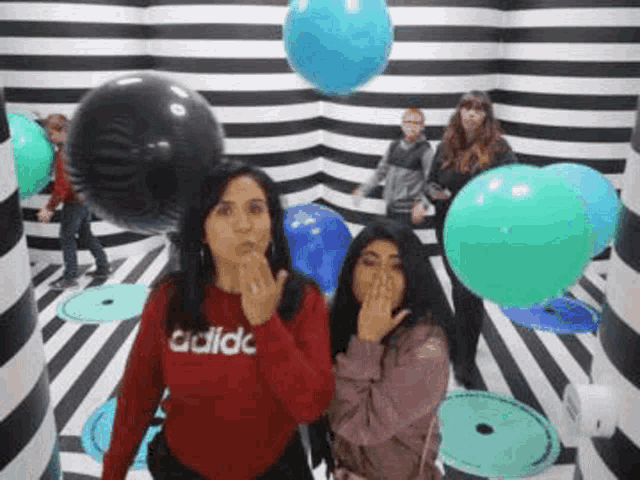 two women are blowing a kiss in a room with balloons and a striped wall .