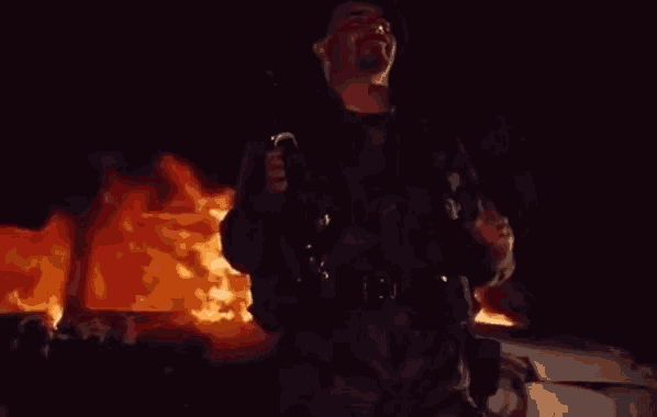 a man holding a gun in front of a burning vehicle