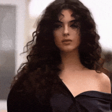 a woman with long curly hair is wearing a black off the shoulder top and looking at the camera .