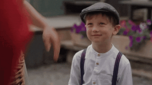 a young boy wearing a flat cap and suspenders is smiling while standing next to a woman .
