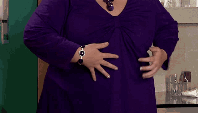 a woman wearing a purple shirt and a silver bracelet is holding her stomach