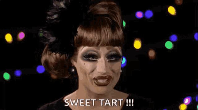 a drag queen is wearing a black dress and feathered headpiece and says sweet tart .