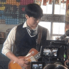 a young man in a vest and tie is playing a guitar in front of a camera .