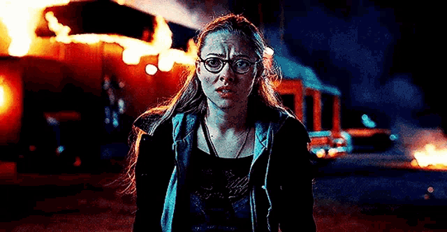 a woman wearing glasses and a black jacket is standing in front of a burning building .