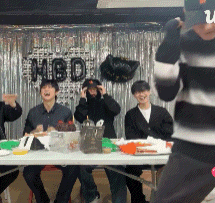 a group of people are sitting around a table with a hbd sign behind them