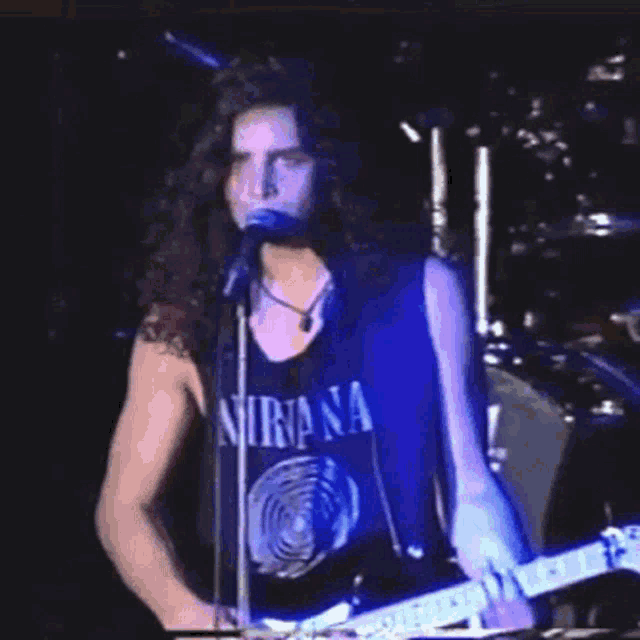 a man in a nirvana shirt is singing into a microphone and playing a guitar .