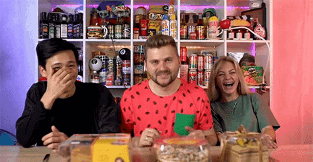 a man in a watermelon shirt is laughing with two other people in front of a shelf full of cans