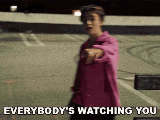 a woman in a pink jacket is pointing at the camera with the words everybody 's watching you below her