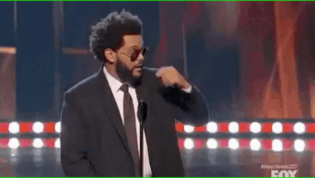 a man wearing sunglasses and a suit is pointing his finger at his nose .
