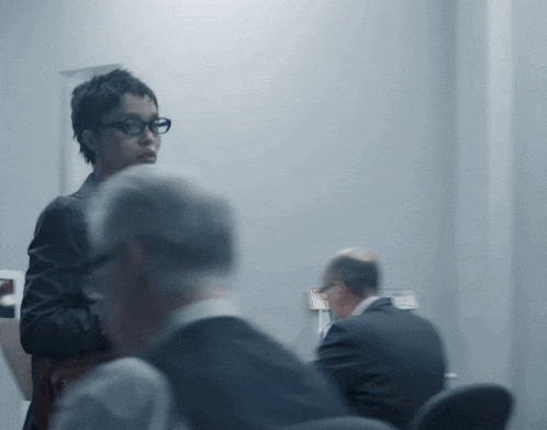 a woman wearing glasses stands in front of a group of men