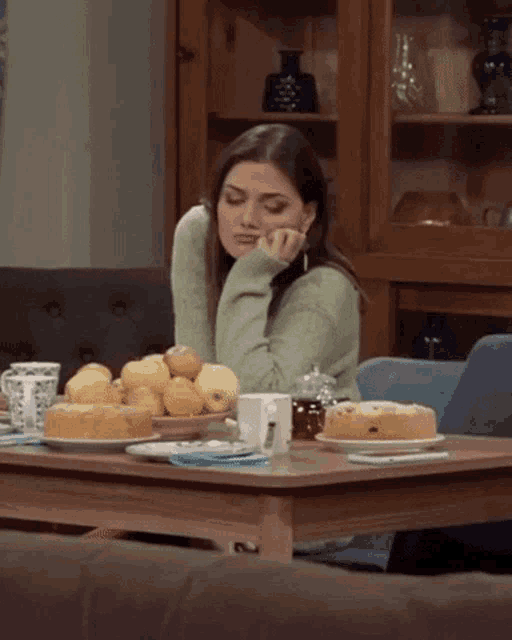 a woman in a green sweater sits at a table with fruit and cakes on it