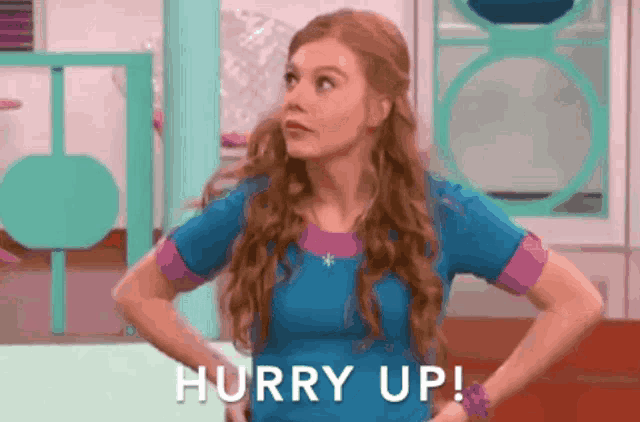 a woman in a blue shirt is standing with her hands on her hips and says hurry up .