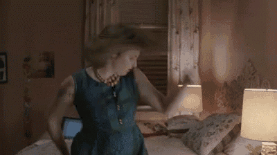 a woman in a blue dress is dancing in a bedroom next to a bed and a lamp .