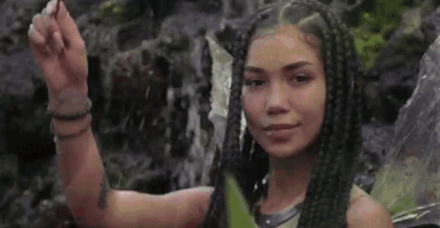 a woman with braids is standing next to a waterfall holding a stick .