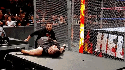 a wrestler is laying on the ground in a cage while a referee stands nearby .