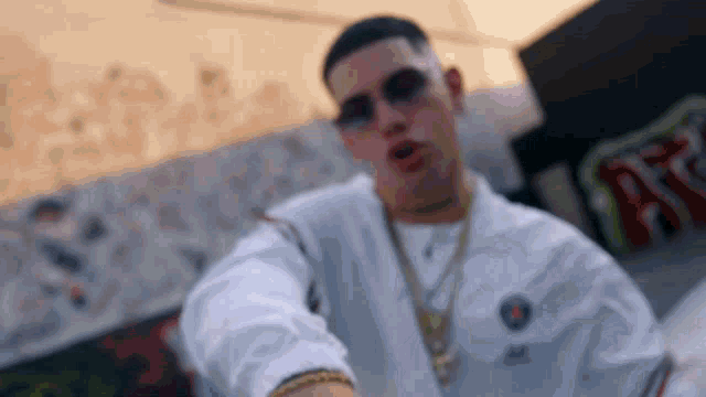 a young man wearing sunglasses and a white shirt is standing in front of a graffiti wall .