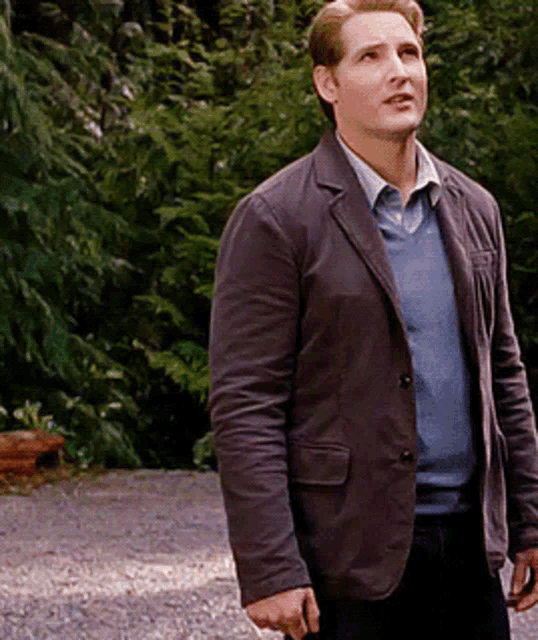 a man wearing a brown jacket and a blue sweater is standing on a gravel road