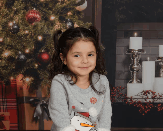 a little girl wearing a sweater with a snowman on it poses in front of a christmas tree and fireplace