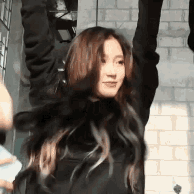 a woman with long hair is standing in front of a brick building with her arms in the air .