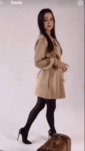 a woman in a trench coat and high heels is walking next to a suitcase .