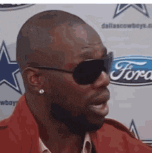 a man wearing sunglasses and earrings stands in front of a ford logo