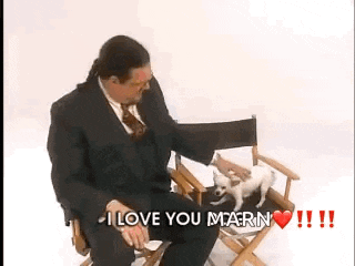 a man in a suit is sitting in a directors chair petting a small white dog .