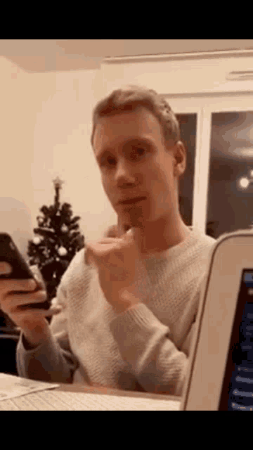 a man is sitting in front of a laptop holding a cell phone .