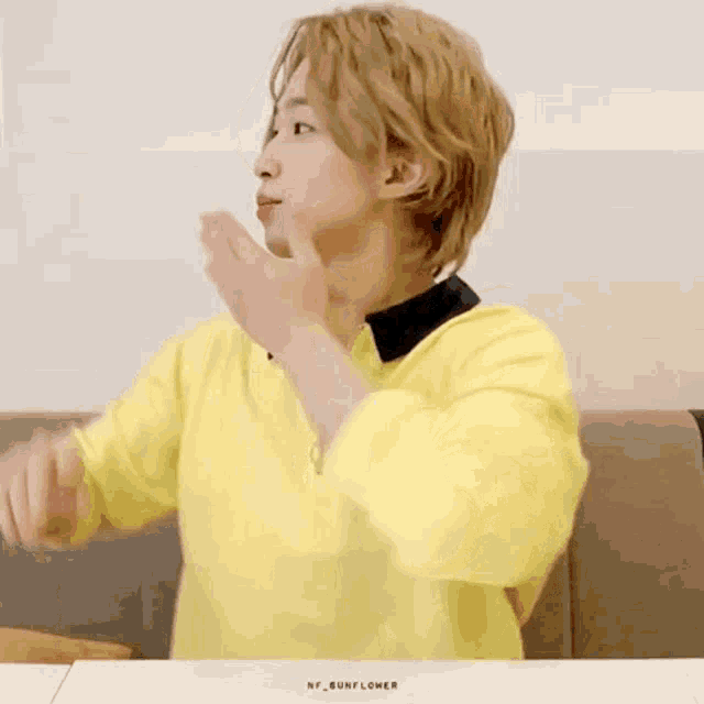 a young man in a yellow sweater is sitting at a table with his hand on his mouth .