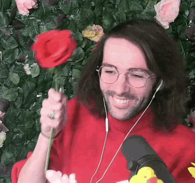 a man wearing glasses and headphones is holding a red rose in front of a wall of flowers .
