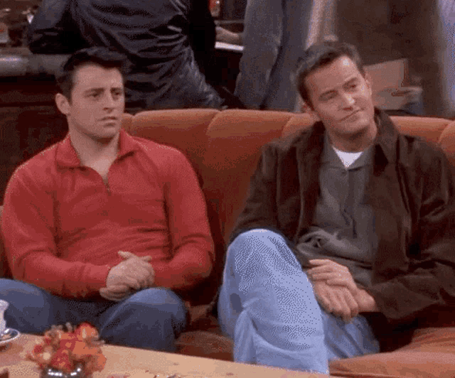 two men are sitting on a couch and one is wearing a red sweater .