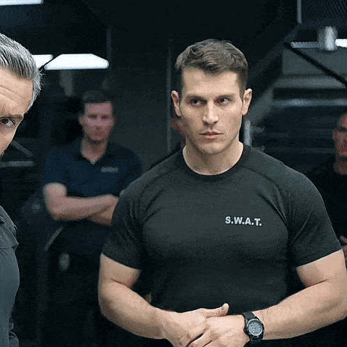 a man wearing a s.w.a.t. t-shirt stands in front of another man